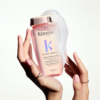 Botella de champu Kerastase Gloss Absolu en manos, con espuma blanca en la parte superior y fondo claro.