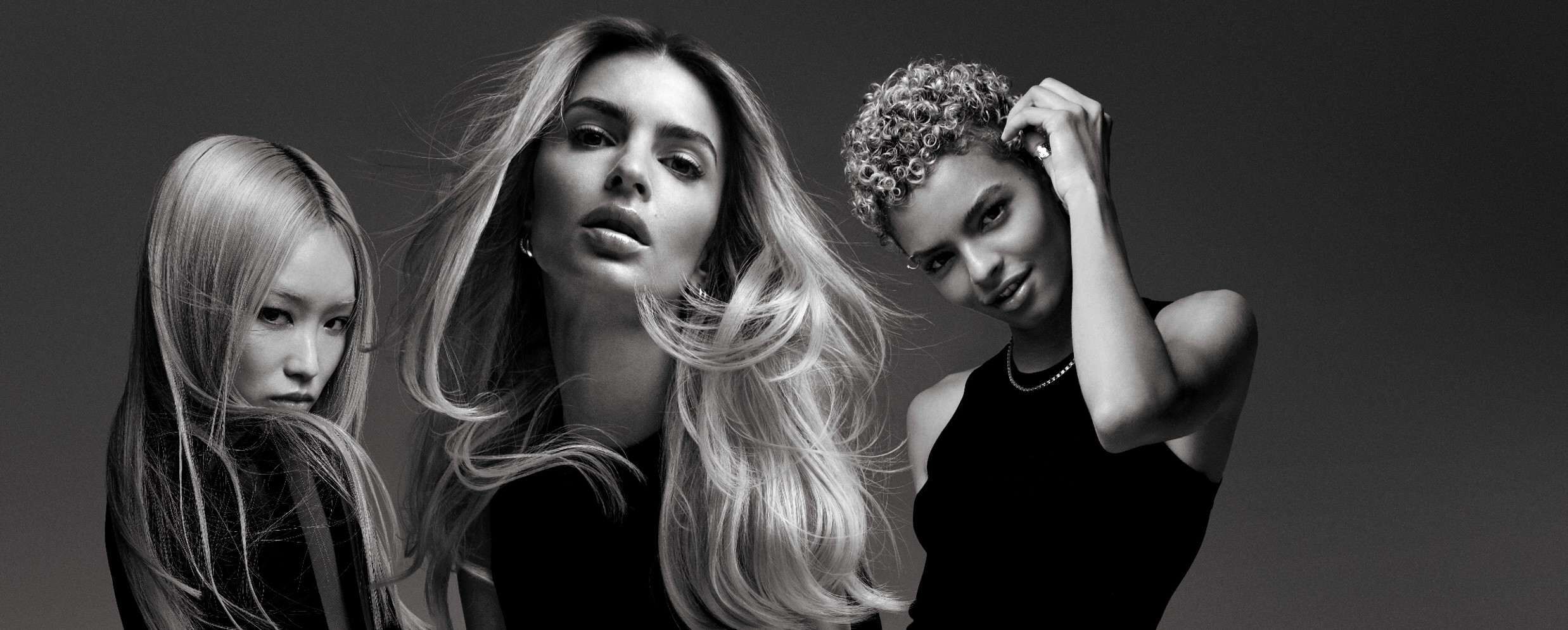 Three women with varying hairstyles pose against a dark background, showcasing stylish black outfits. Their expressions and hair create a dynamic, artistic composition.