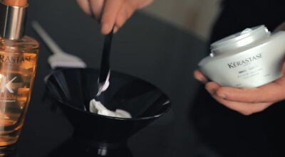 A hand holds a black mixing bowl containing a white cream, with a black spatula for mixing; a bottle of hair oil is visible in the background.