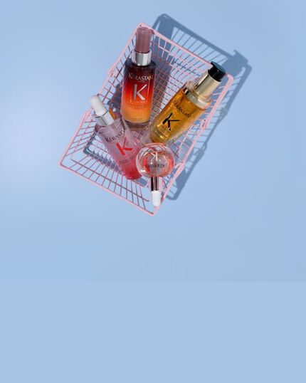 Cuatro productos capilares Kerastase en mini-carritos rosas sobre fondo azul: el Serum Noche 8H Nutritive, el Aceite Original Elixir Ultime, el Serum Anticaida Fortificante de Genesis y las Glaze Drops.