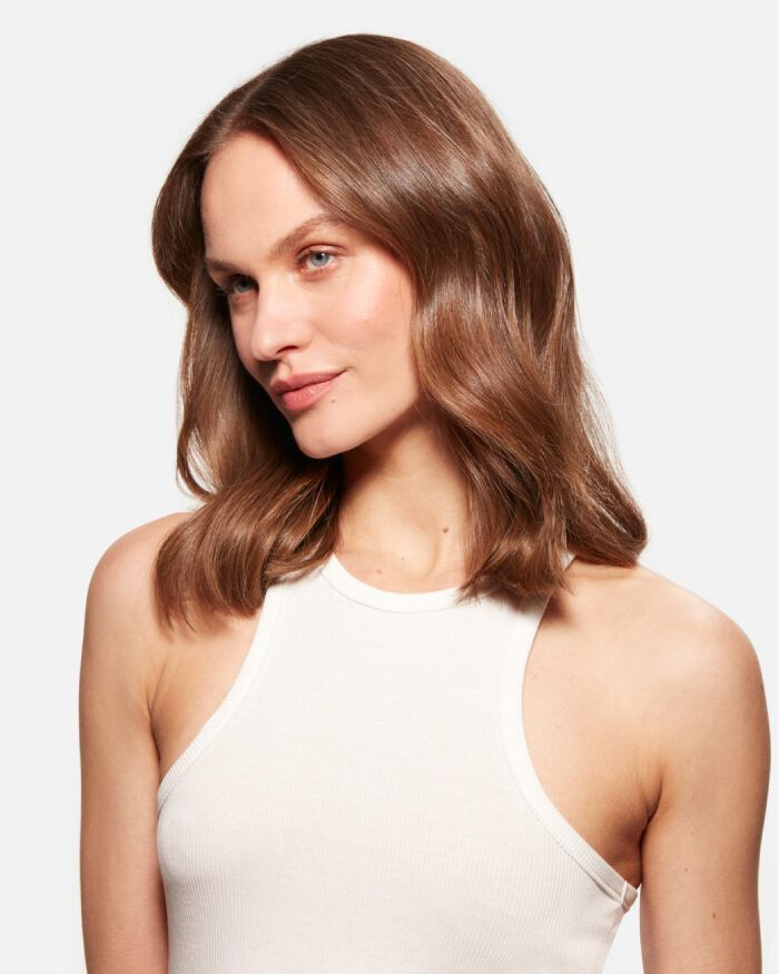 Mujer con cabello castano y ondulado, mirando hacia un lado, vestida con una camiseta sin mangas blanca.