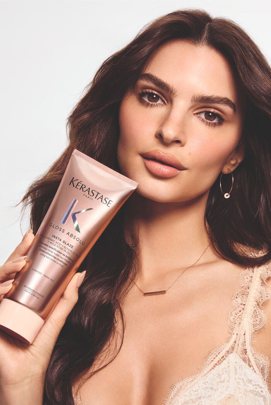 A woman with long, wavy hair holds a tube of Kerastase Gloss Absolu hair product close to her face, showcasing her radiant skin and neutral makeup against a light background.