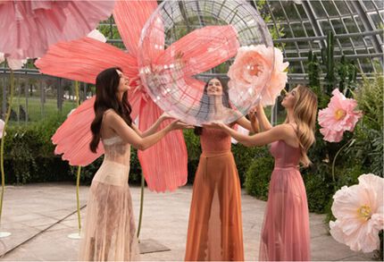 Tres mujeres con vestidos fluidos interactuando con burbujas transparentes y flores gigantes rosas en un entorno exuberante.