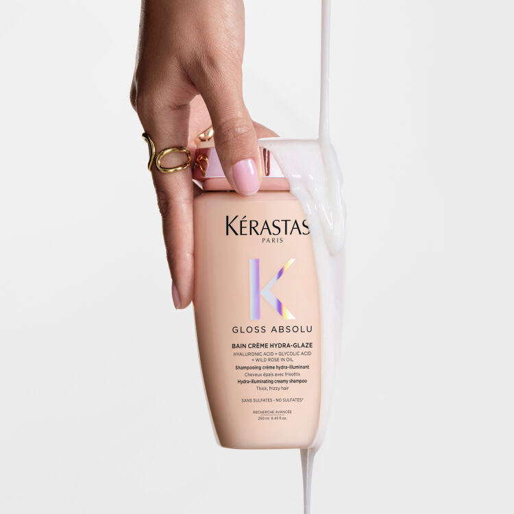 Mano sosteniendo una botella de champu Kerastase Gloss Absolu con un liquido blanco cremoso cayendo sobre ella.