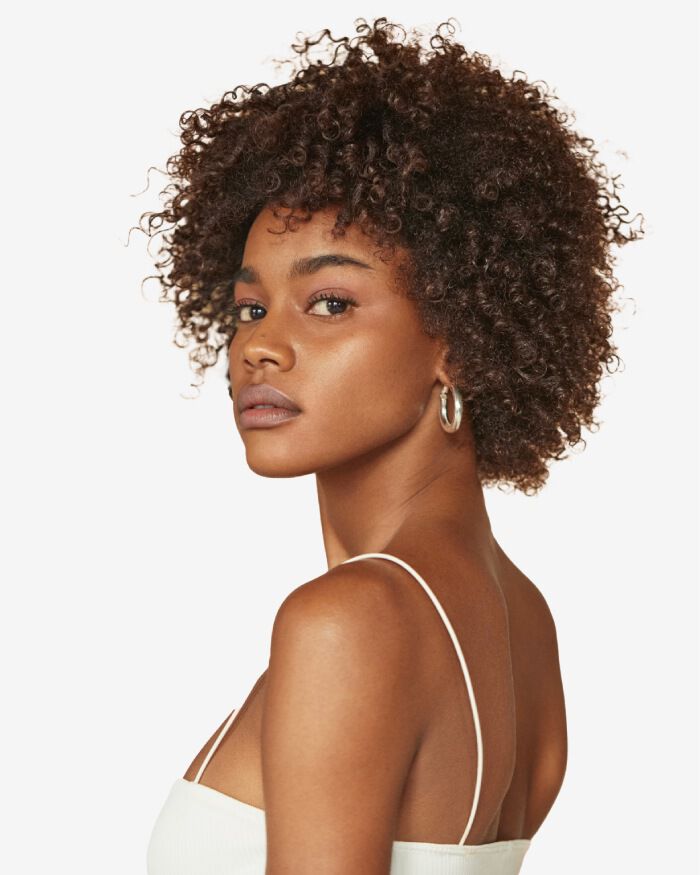 Mujer con cabello rizado y suelto, mirando hacia un lado, con una blusa sin mangas y aretes grandes.