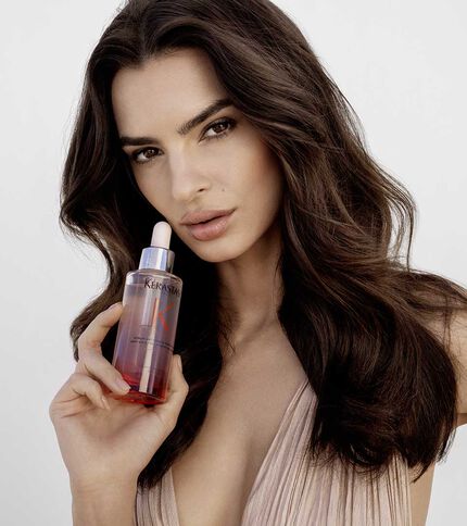A model with long, wavy hair holds a bottle of hair serum close to her face, wearing a subtle, natural makeup look against a plain background.