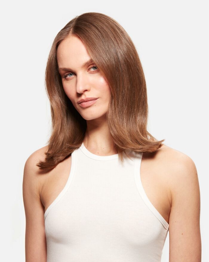 Una persona con cabello lacio y brillante, viste una camiseta de tirantes blanca y mira hacia la camara con una expresion segura.