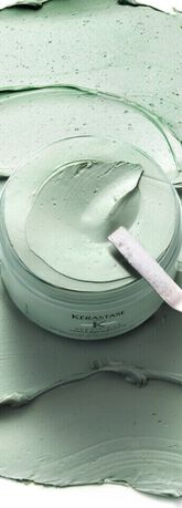 A jar of green hair mask surrounded by a smooth, textured application of the same product, with a metal spatula resting on top.