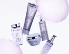 A collection of Kerastase hair care products displayed elegantly on a iridescent background. The products include a shampoo, conditioner, a treatment, and a serum, all presented in sleek packaging with a lavender theme.