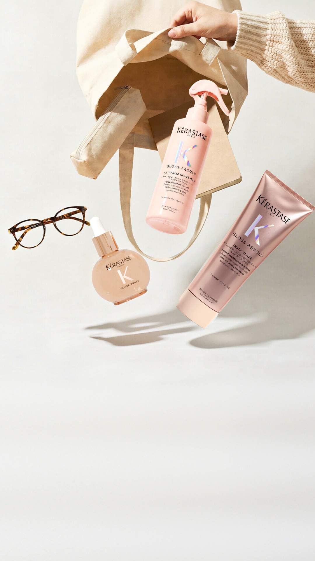 Tres frascos de productos capilares Kerastase (Elixir Ultime, Genesis y Nutritive) estan dispuestos sobre una superficie blanca junto a una camara de fotos vintage y varias peonias blancas con hojas verdes. El frasco dorado Elixir Ultime esta a la izquierda con una bomba negra, el frasco rosa Genesis con un gotero plateado esta en el centro, y el frasco degradado rojo-naranja Nutritive con un gotero plateado esta a la derecha. La camara plateada con su correa negra se encuentra a la izquierda de los productos.