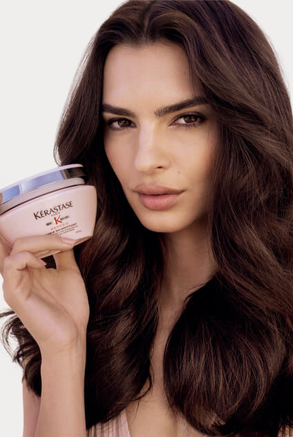 Mujer sosteniendo un tarro rosa de la mascarilla Kerastase Genesis Masque Reconstituant, presentando el producto.