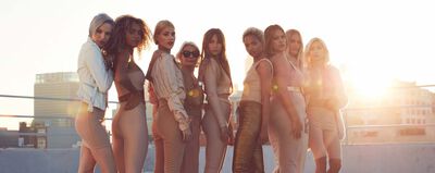 A diverse group of nine women poses confidently in fashionable, form-fitting outfits against a sunset backdrop, showcasing a blend of styles and a sense of empowerment.