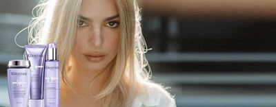 A model with long, light blonde hair is featured prominently alongside Kerastase hair care products displayed in the foreground, including shampoo, conditioner, and a treatment. The background has a soft, blurred effect, creating a focus on the model and the products.