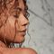 A woman with curly hair holds a jar of hair treatment while standing in a shower, surrounded by marble tiles.
