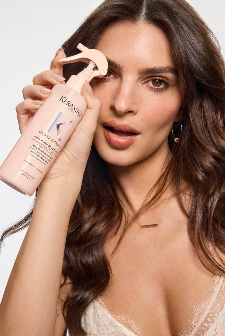 A woman with long, wavy hair holds up a spray bottle of Kerastase hair product close to her face, smiling gently. She wears natural makeup and a delicate necklace.