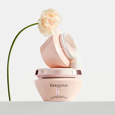 Dos tarros de mascarilla capilar Kerastase Gloss Absolu con textura blanca cremosa saliendo de uno y un clavel rosa palido.