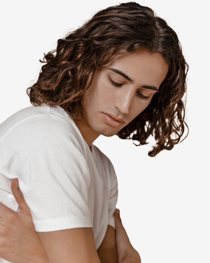 Un joven con cabello rizado y largo, vestia una camiseta blanca, mirando hacia abajo con una expresion tranquila.