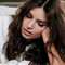 A woman with long, wavy hair is resting her head on her hand, looking down thoughtfully. She is dressed in a white shirt and appears relaxed in a cozy setting.