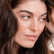 A close-up portrait of a woman with long, wavy brown hair, looking thoughtfully to the side with a soft expression.
