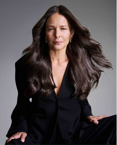 Retrato de una mujer madura con cabello largo castano y voluminoso, vistiendo un blazer negro sobre fondo gris.