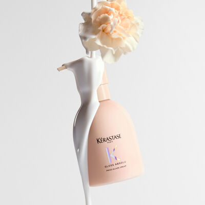 Botella de crema Kerastase Gloss Absolu Frizz-Glaze con un liquido blanco cremoso cayendo sobre ella y una flor de clavel palido en la parte superior.