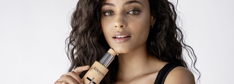 Una mujer con cabello rizado y oscuro sostiene un frasco de base de maquillaje de la marca Kerastase, luciendo una expresion suave y natural.