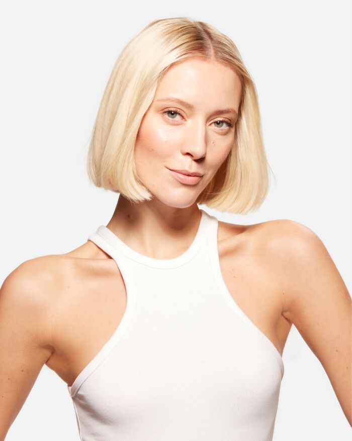 Un modelo con cabello rubio y corto, luciendo una camiseta sin mangas blanca, sonrie suavemente.