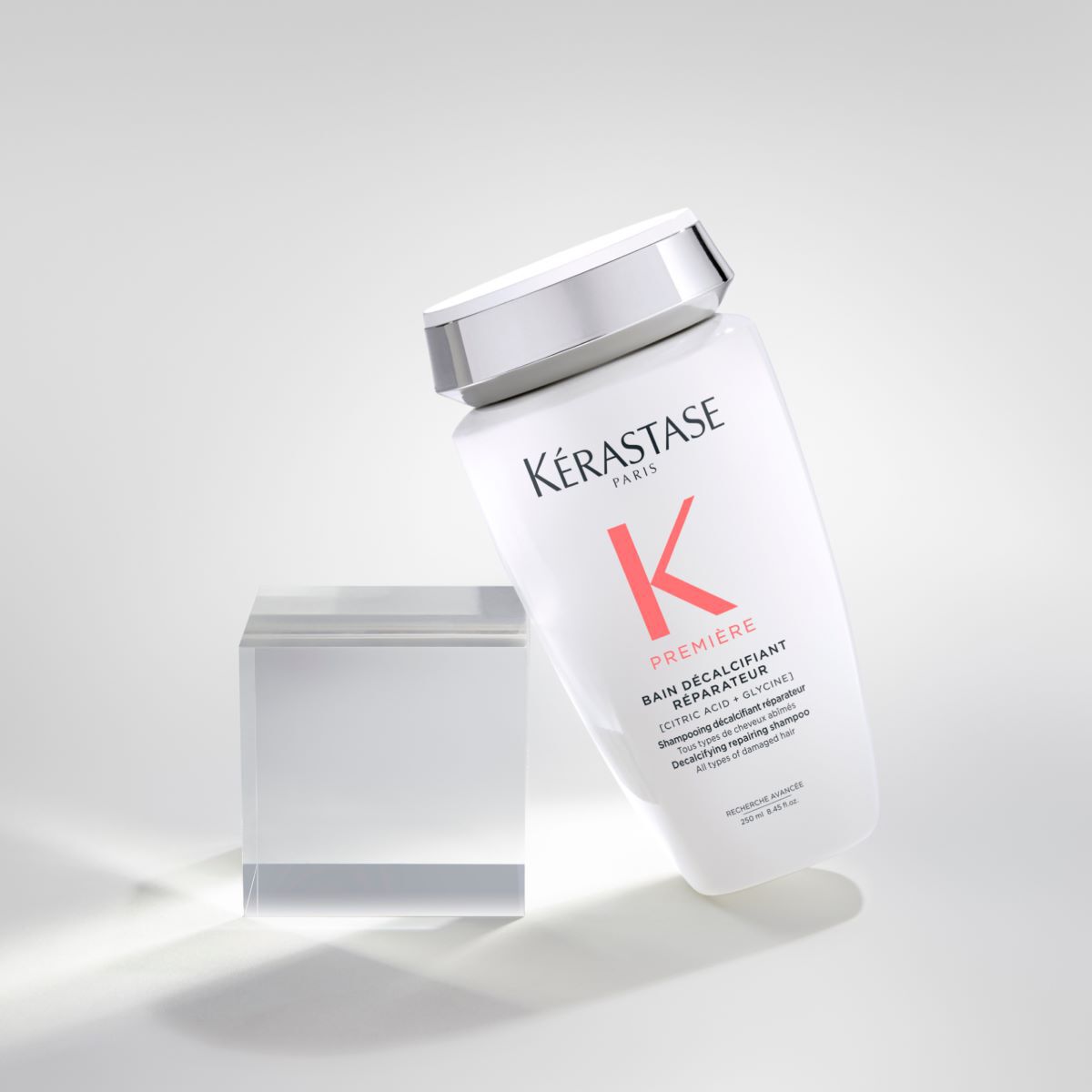 Botella de champu Kerastase en posicion inclinada sobre un fondo claro, junto a un bloque de acrilico transparente.