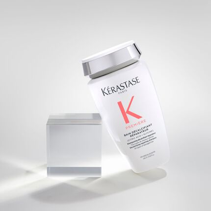 Botella de champu Kerastase en posicion inclinada sobre un fondo claro, junto a un bloque de acrilico transparente.