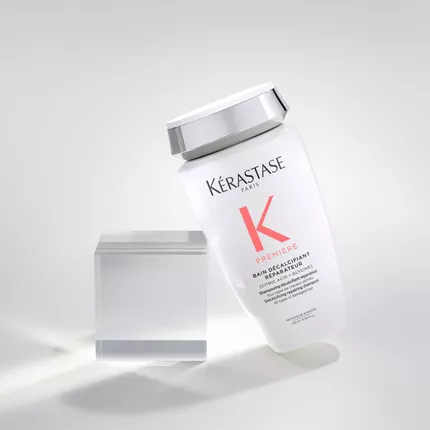 Botella de champu Kerastase en posicion inclinada sobre un fondo claro, junto a un bloque de acrilico transparente.