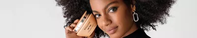 A young woman with curly hair holds a gold hair care product close to her face, smiling confidently against a white background.