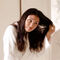 A woman in a white bathrobe is brushing her long hair while looking to the side. The background features a light-colored wall and a small electrical outlet.