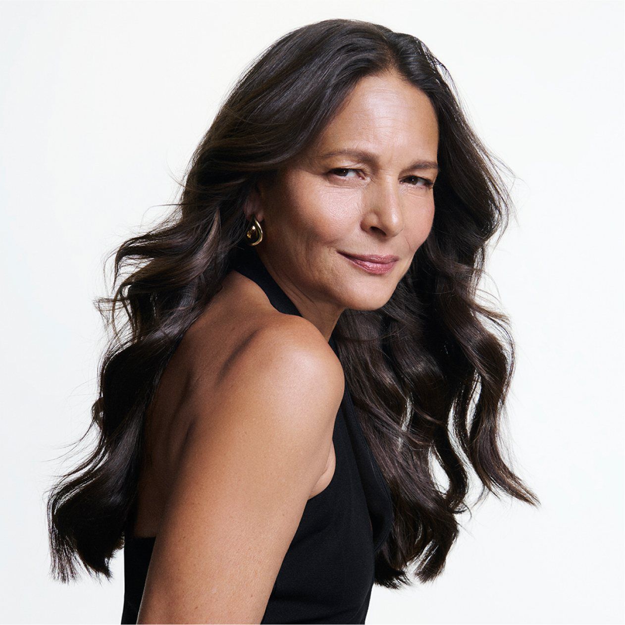 Retrato de una mujer madura con cabello largo, oscuro y ondulado, posando con una sonrisa segura sobre fondo blanco.