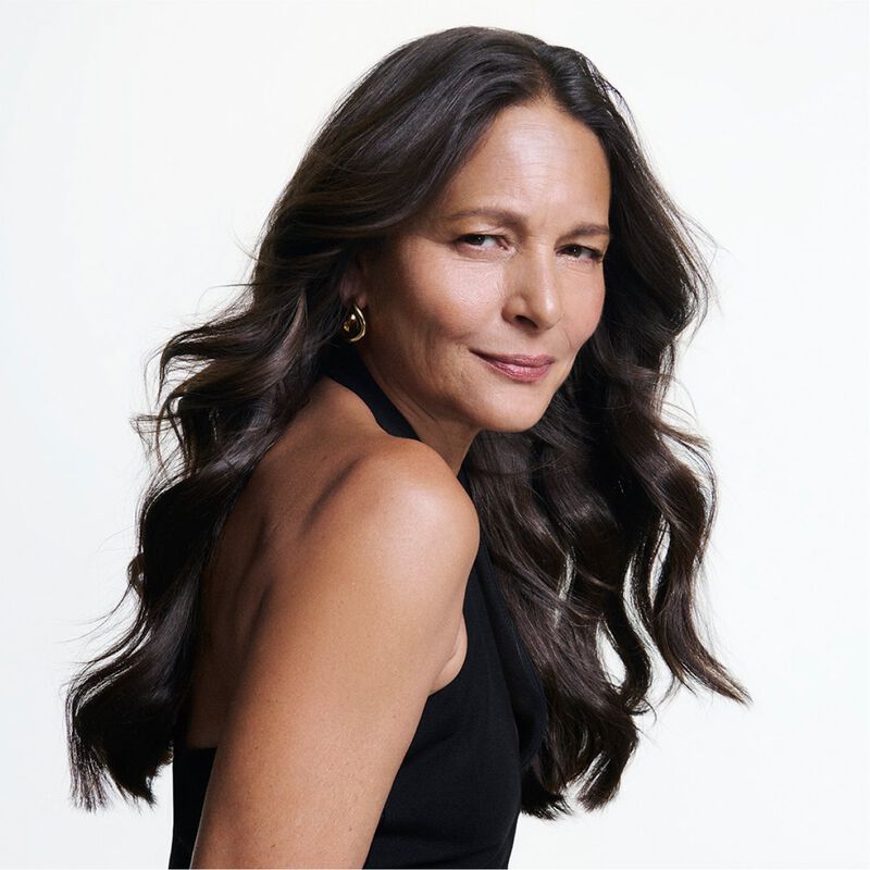 Retrato de una mujer madura con cabello largo, oscuro y ondulado, posando con una sonrisa segura sobre fondo blanco.