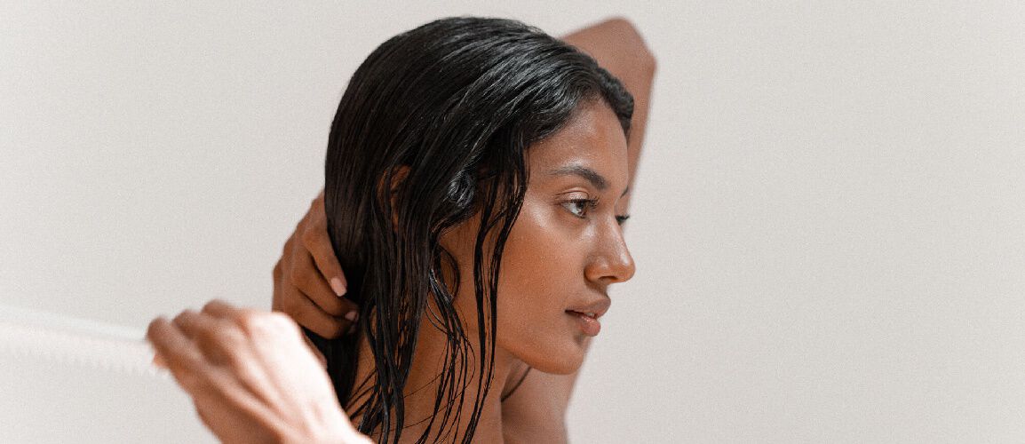 A profile view of a woman with long, wet hair, gently combing her hair while her hand is positioned behind her head. The background is neutral and minimalistic.