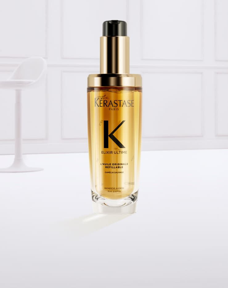 Frasco de suero Kerastase Elixir Ultime rodeado de gotas de aceite dorado sobre un fondo claro.