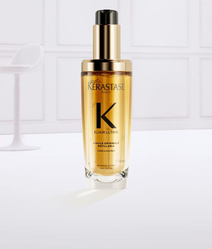 Frasco de suero Kerastase Elixir Ultime rodeado de gotas de aceite dorado sobre un fondo claro.