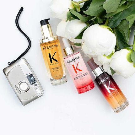 Tres frascos de productos capilares Kerastase (Elixir Ultime, Genesis y Nutritive) estan dispuestos sobre una superficie blanca junto a una camara de fotos vintage y varias peonias blancas con hojas verdes. El frasco dorado Elixir Ultime esta a la izquierda con una bomba negra, el frasco rosa Genesis con un gotero plateado esta en el centro, y el frasco degradado rojo-naranja Nutritive con un gotero plateado esta a la derecha. La camara plateada con su correa negra se encuentra a la izquierda de los productos.