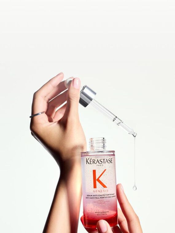 Primer plano de manos sosteniendo una botella de Kerastase Genesis Serum Fortificante, con un cuentagotas del que esta a punto de caer una gota de producto.
