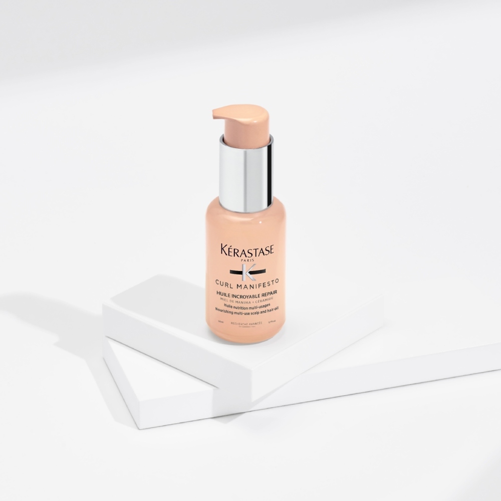 El aceite capilar Curl Manifesto Huile Sublime Repair se presenta sobre un fondo gris con cubos transparentes. Hidrata y da forma a los rizos a la vez que actúa contra el encrespamiento y la rotura.