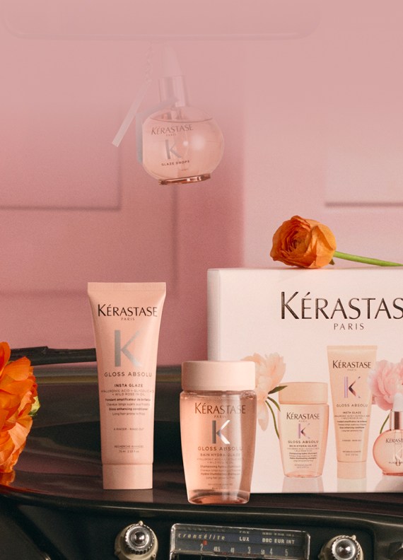Imagen de productos capilares Kerastase arreglados como un regalo, que incluyen varios frascos dorados, una vela perfumada y una bolsa blanca de rizo, todo presentado en un empaque festivo blanco y dorado sobre un fondo blanco y luminoso. El conjunto evoca lujo y elegancia.