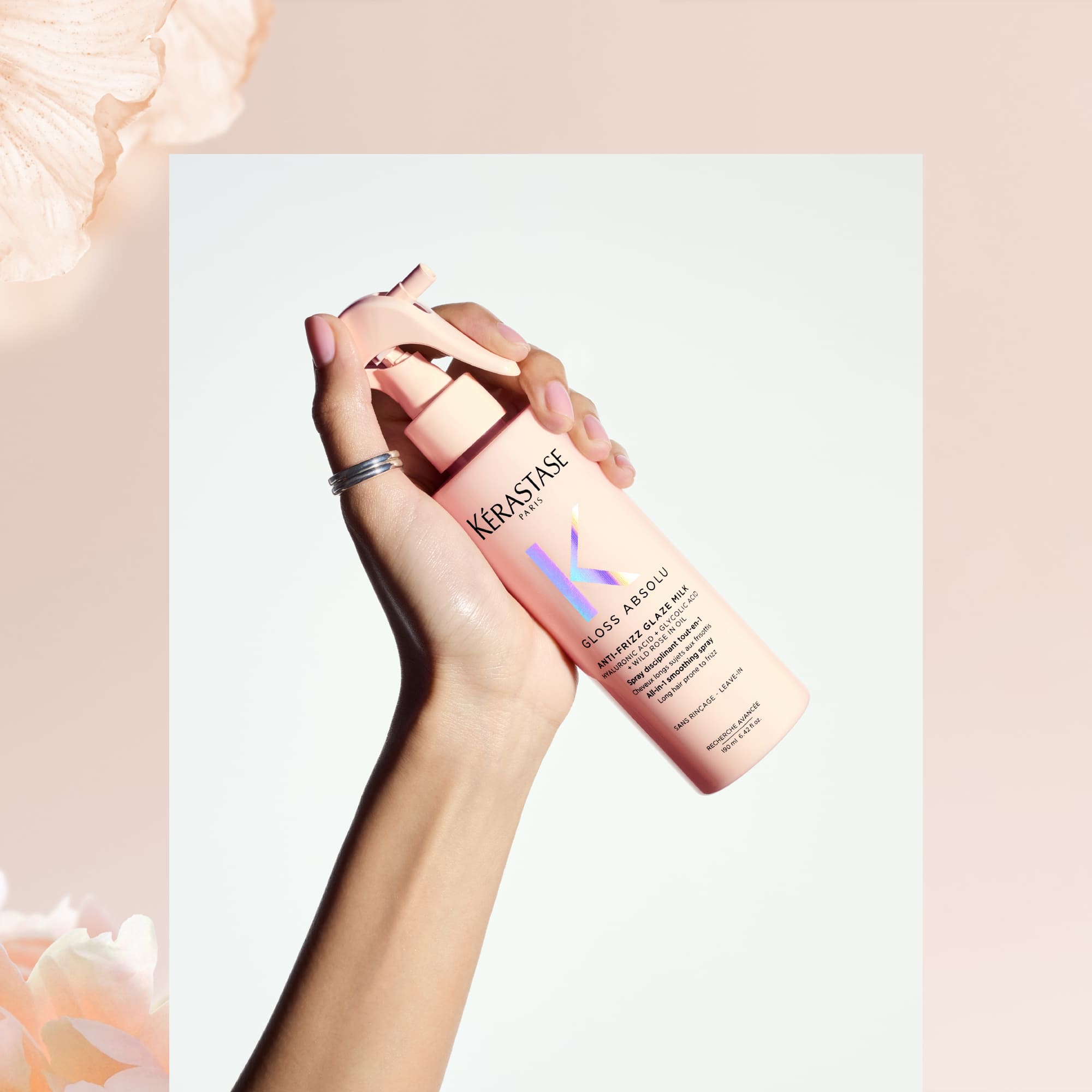 Frasco de spray de Kerastase en mano, fondo minimalista con flores suaves. Producto para el cuidado del cabello.