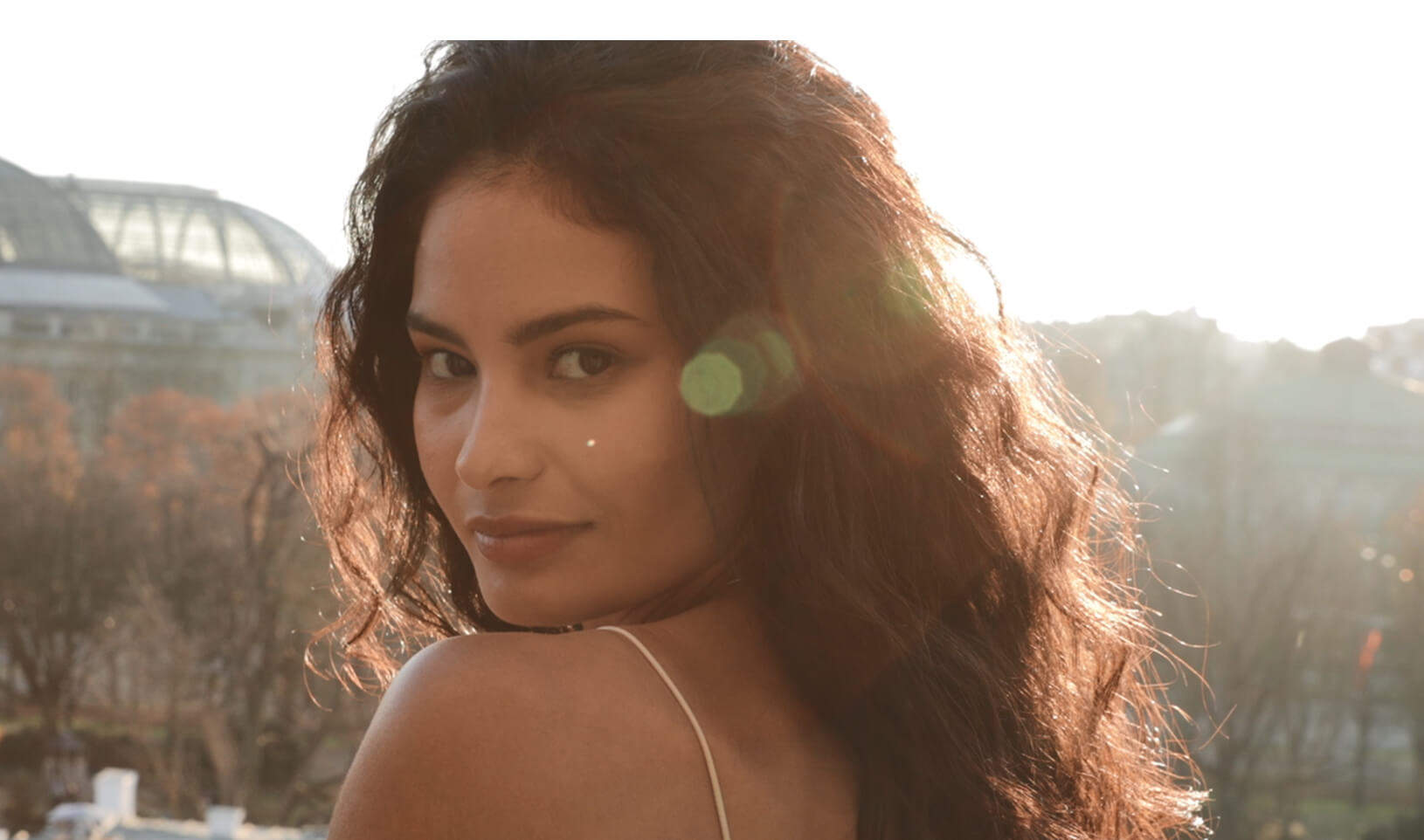 Mujer de cabello rizado y suelto, mirando hacia atrás con una suave luz solar de fondo en un entorno urbano.