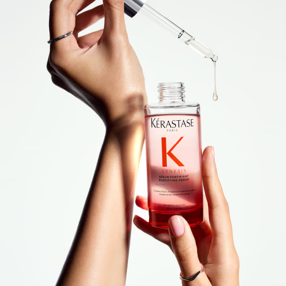 Primer plano de manos sosteniendo una botella de Kérastase Genesis Sérum Fortificante, con un cuentagotas del que está a punto de caer una gota de producto.