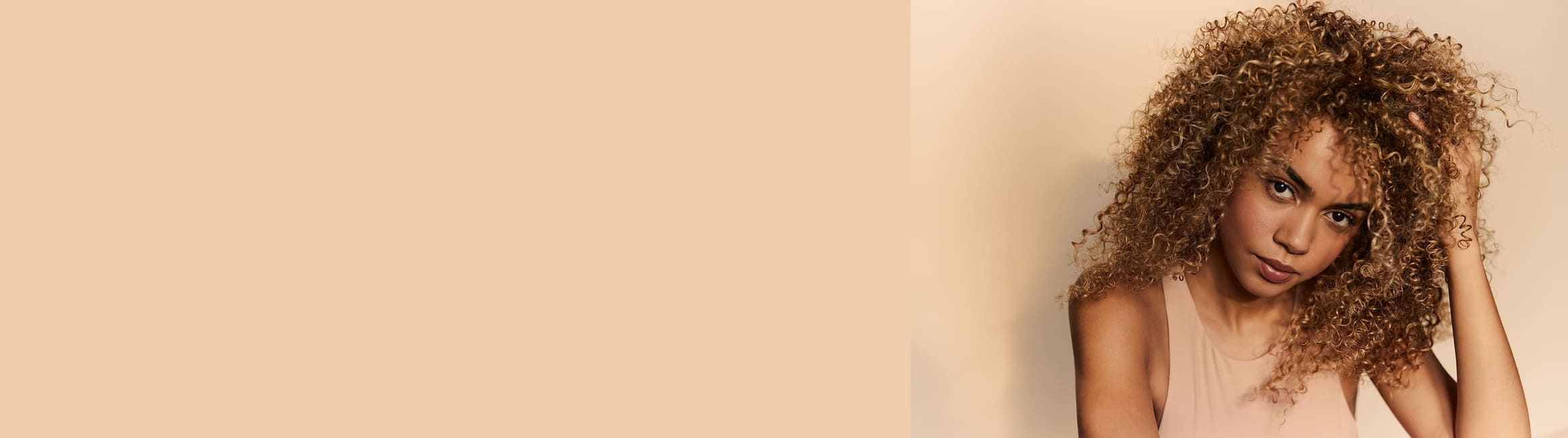 Una mujer con cabello rubio rizado y voluminoso posa frente a un fondo de color crema. Mira a la cámara con una expresión pensativa, con la mano ligeramente apoyada en su cabello.